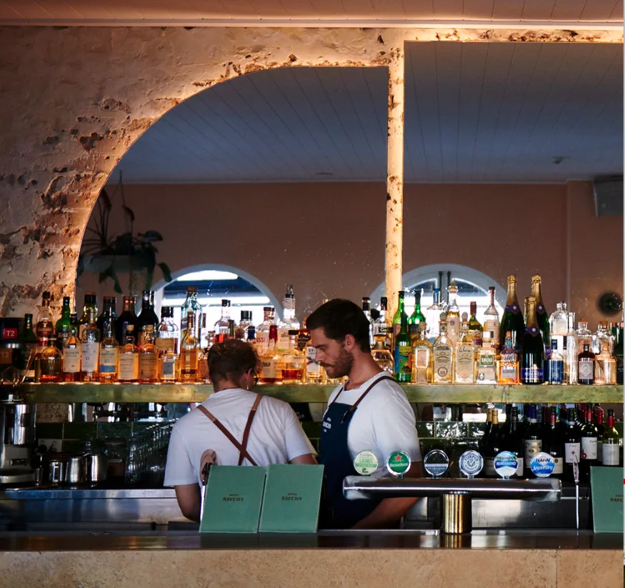 Two bartenders behind the long brass bar at Hotel Ravesis, under the sandstone arches